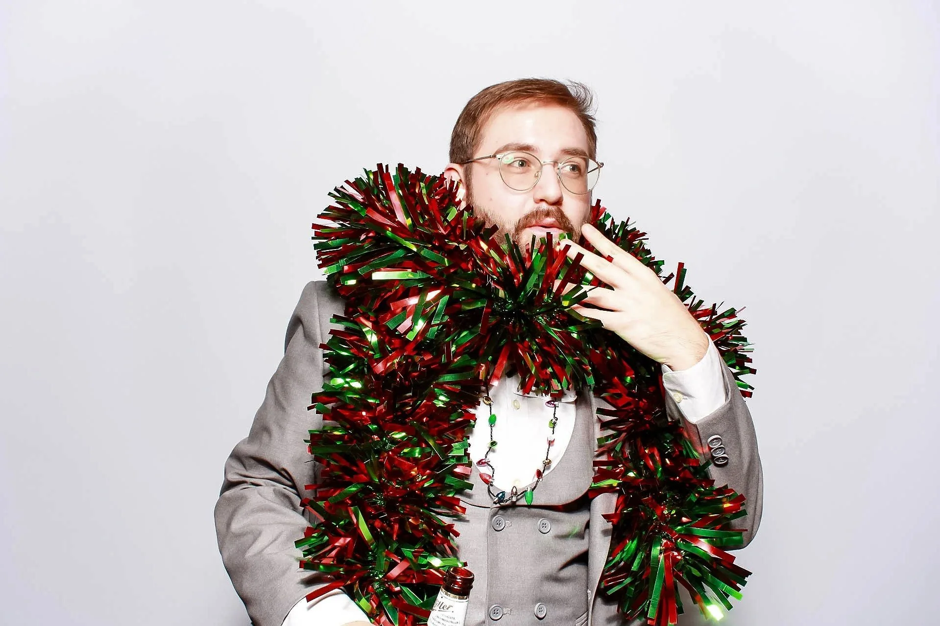 A man with glasses and a beard wearing a gray suit is surrounded by red and green tinsel, holding a bottle of alcohol, and posing against a plain white background.