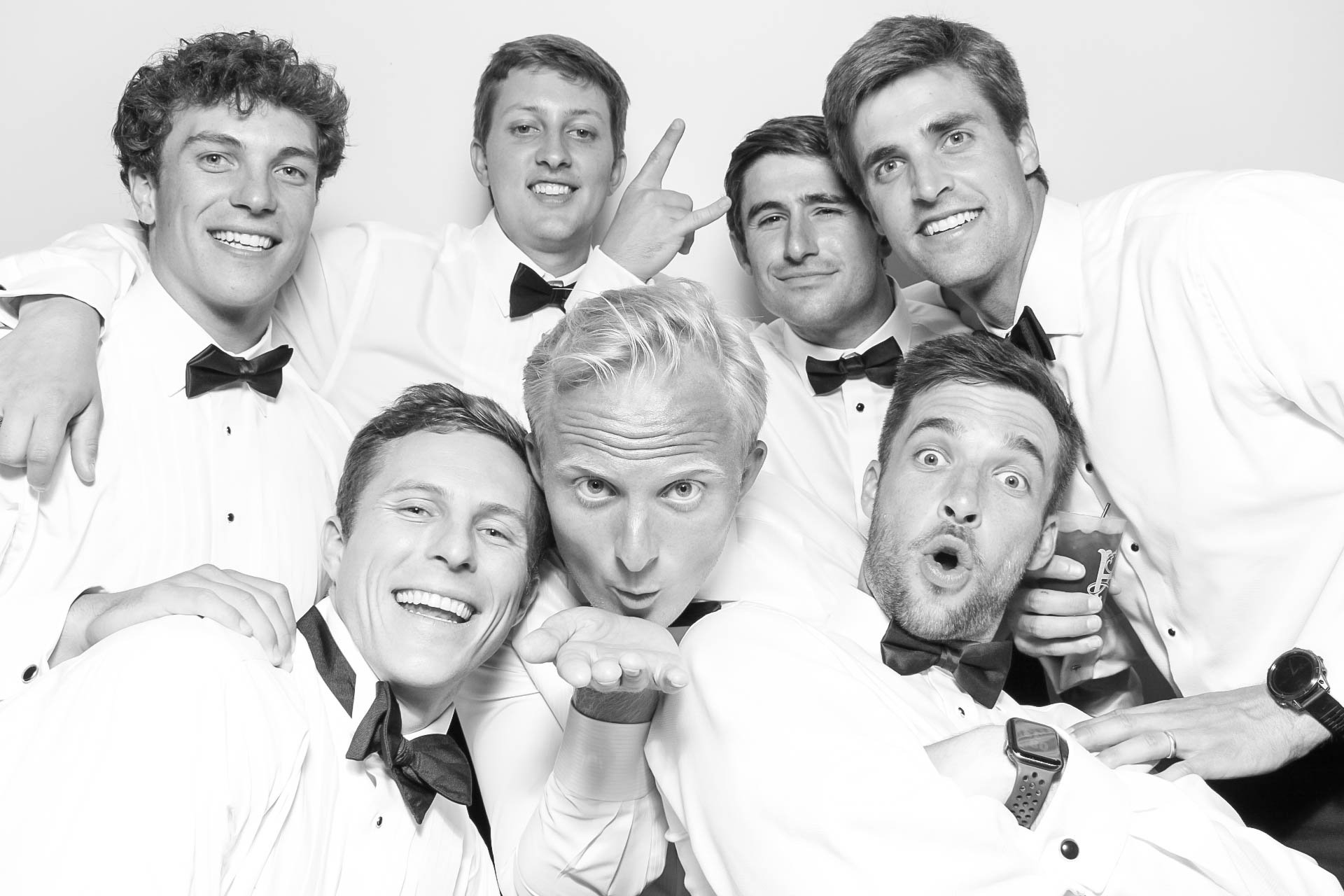 Group of young men dressed in tuxedos and bow ties, smiling and posing closely for a photo in a black-and-white studio setting.