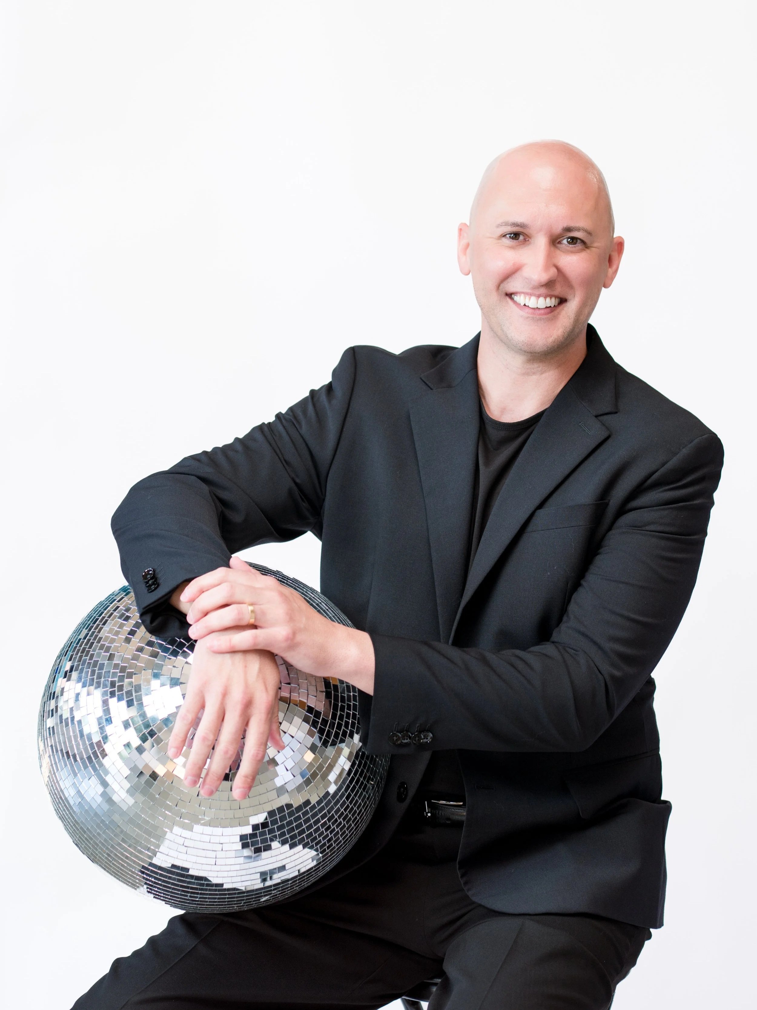 A smiling, bald man in a black blazer holding a large disco ball while posing against a plain white background.