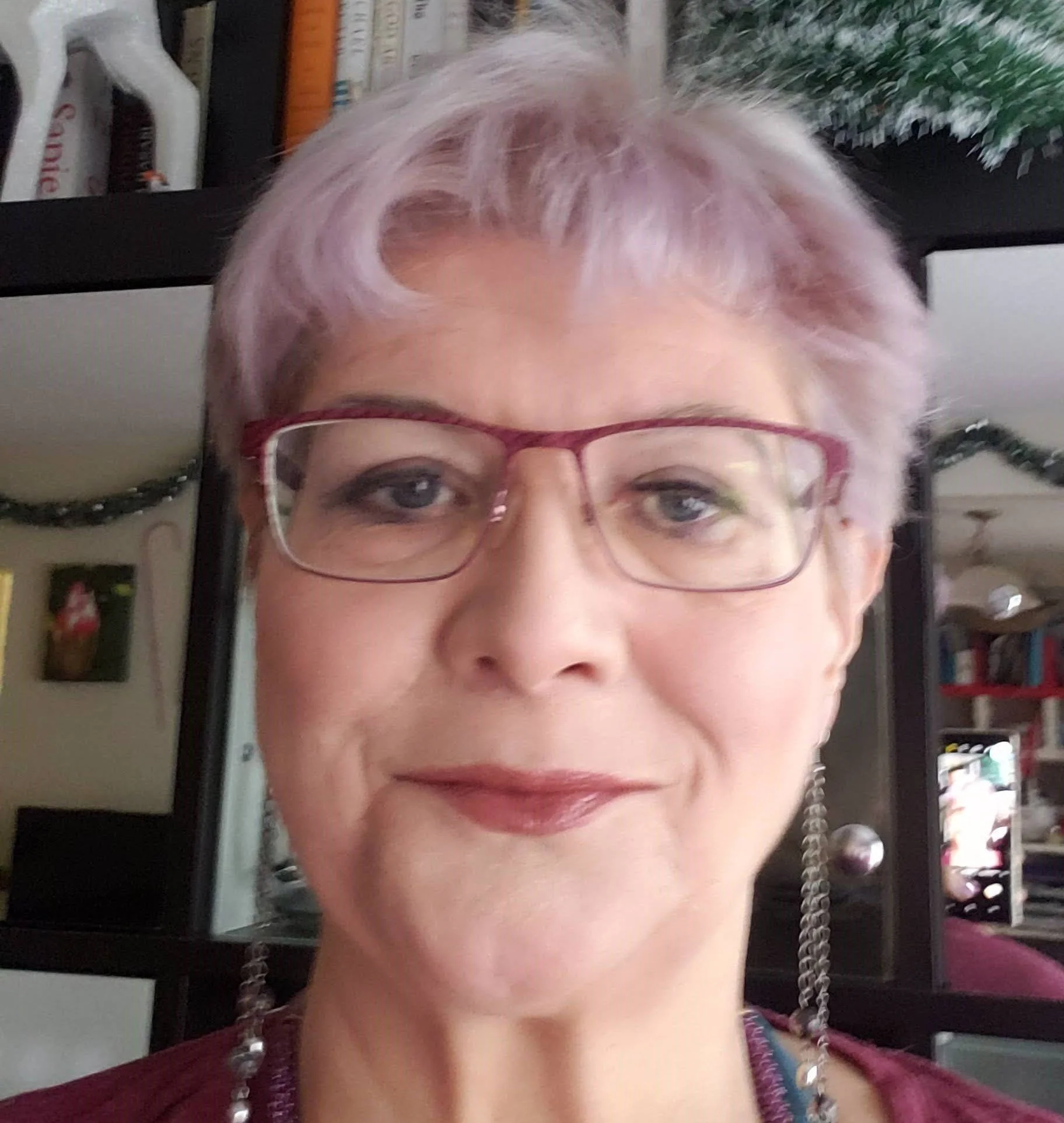 Close-up of a woman with short, light-colored hair and red glasses, smiling slightly, with books and plants visible in the background.