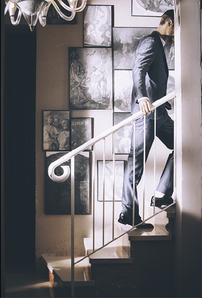 Modello in completo scuro mentre sale una scala in un interno decorato con opere d’arte, editoriale moda uomo per Santaniello fotografato da Paolo Esposito.