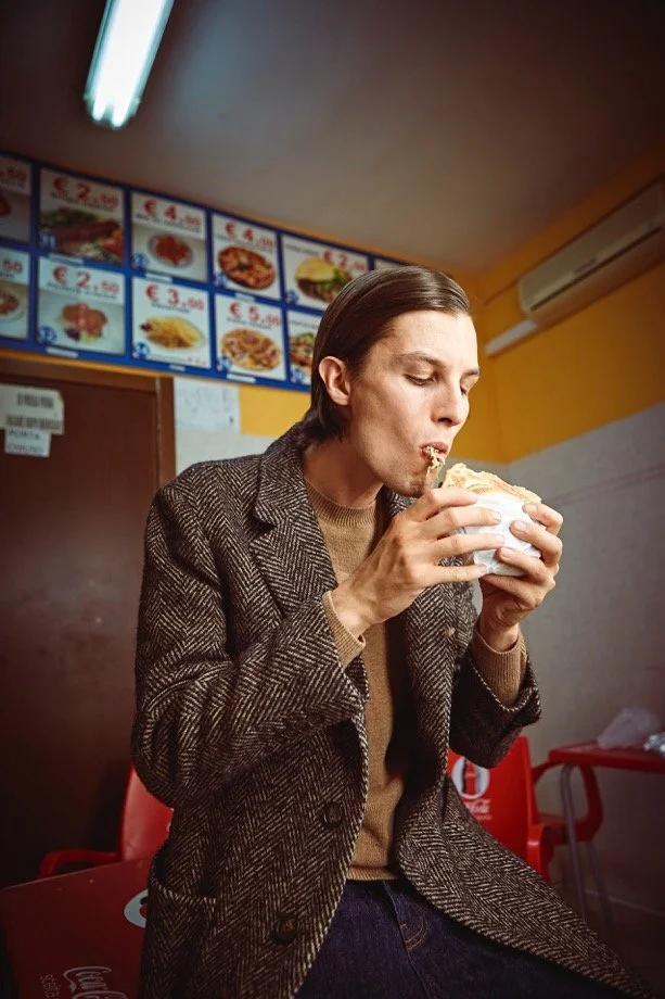 Modello in cappotto spinato mentre mangia un panino in un locale, editoriale moda uomo per Biagio Santaniello.