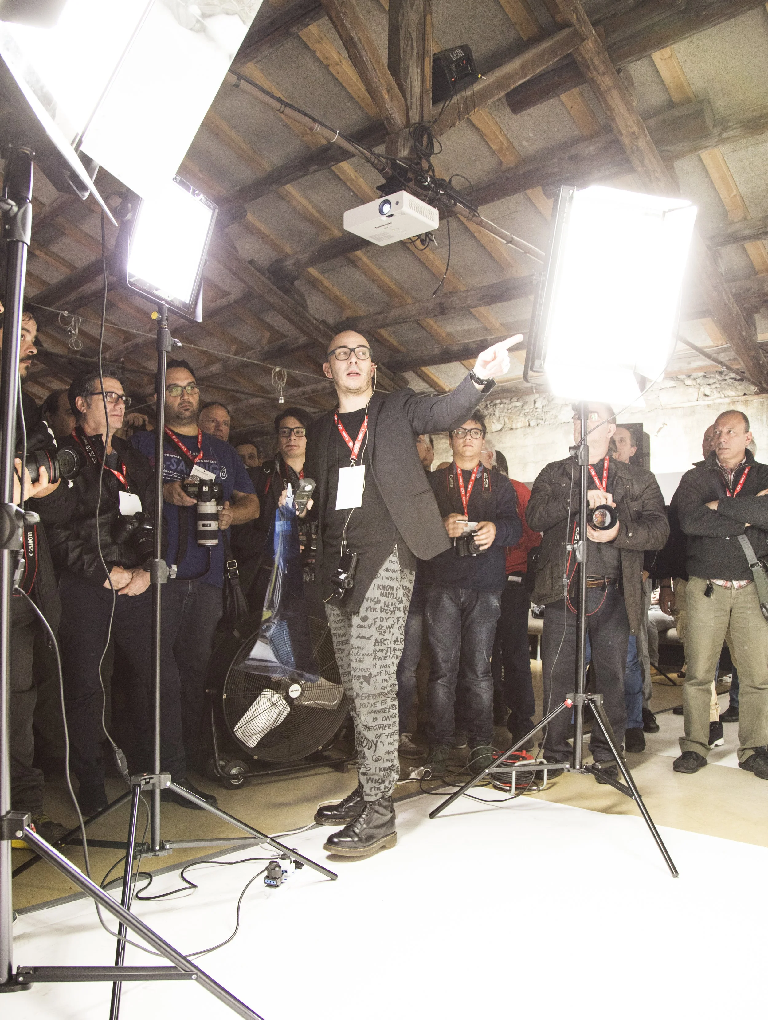 Un uomo con occhiali neri e una giacca nera sta spiegando qualcosa a un gruppo di persone durante una presentazione in un ambiente industriale. Ci sono luci intense, tre persone con fotocamere e altri con badge attorno al collo.