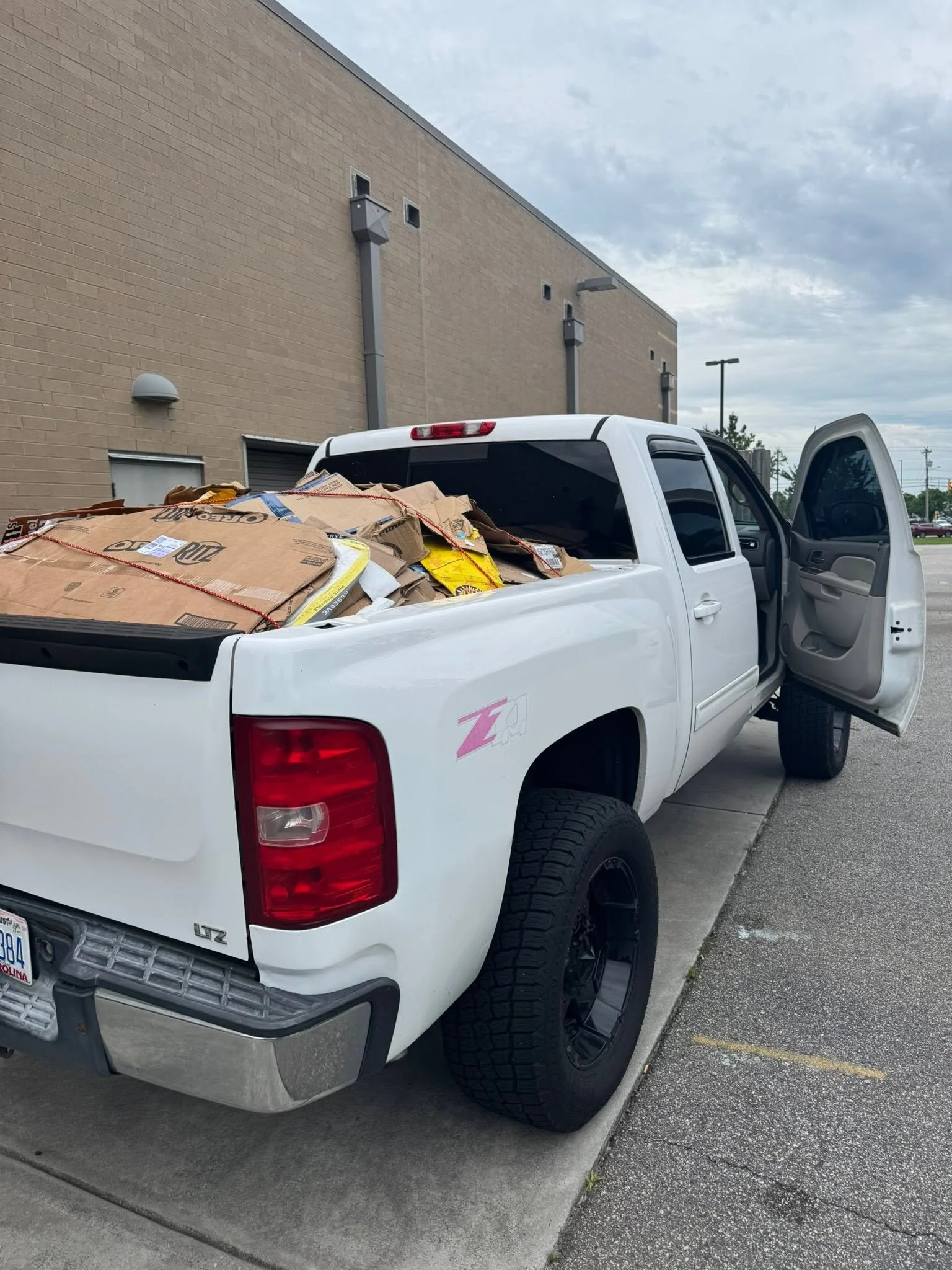 A white pickup truck parked next to a sidewalk outside a beige brick building, with boxes stacked in the truck bed and the passenger door open.