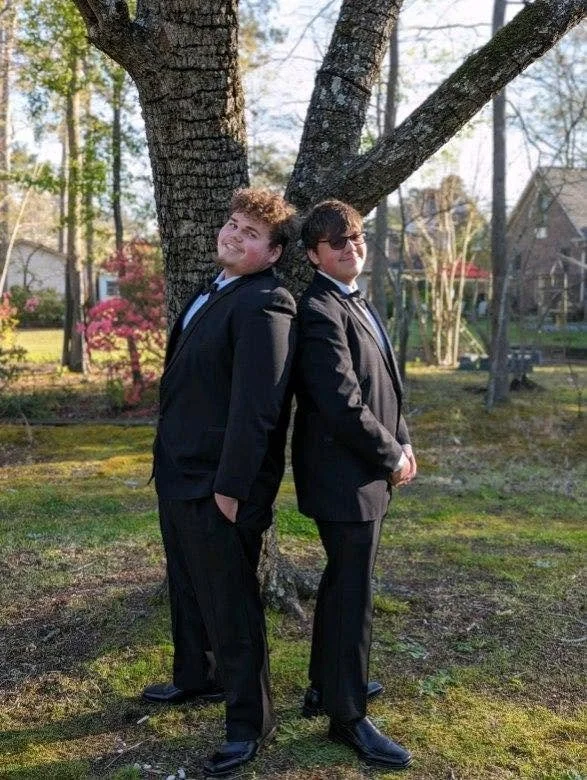 Two young men in black suits and sunglasses stand back to back, leaning against a tree in a garden with flowering bushes and residential houses in the background.