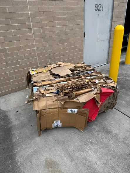 Pile of flattened cardboard boxes leaning against a wall near a door with the number 821, next to yellow safety bollards on a concrete ground.