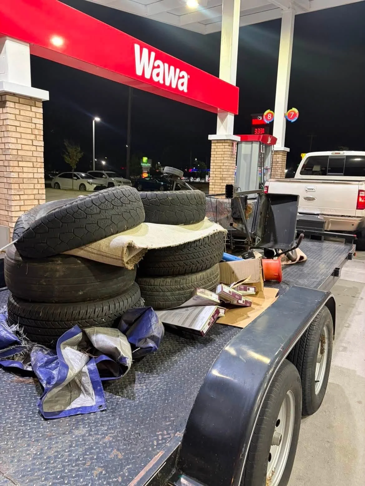 A flatbed trailer loaded with used car tires, some stacked and others scattered, at a Wawa gas station at night.