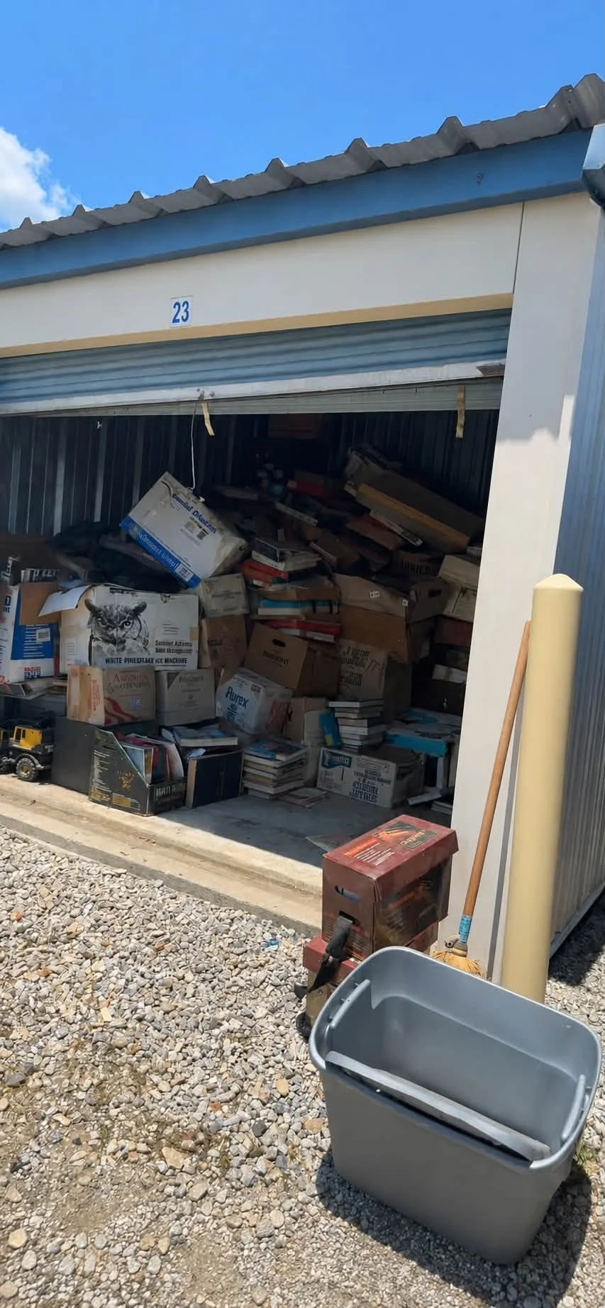 Open storage unit filled with boxes, books, and miscellaneous items, with a gray trash bin and cleaning tools outside on gravel ground.