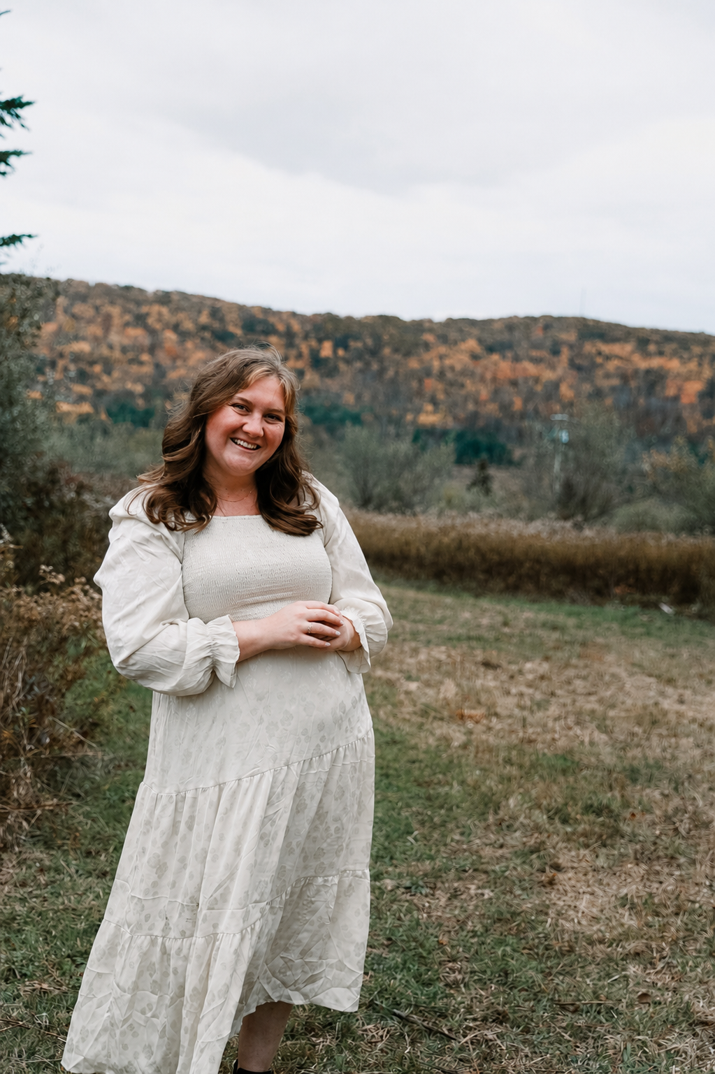A smiling woman in a long white dress standing outdoors in a natural setting with hills and trees in autumn colors in the background.