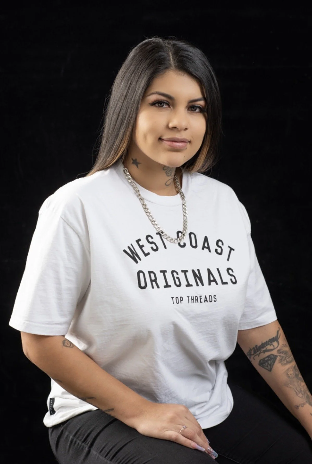 A young woman with long dark hair, tattoos on her arms and neck, wearing a white t-shirt with black text and a silver chain necklace, sitting against a black background.