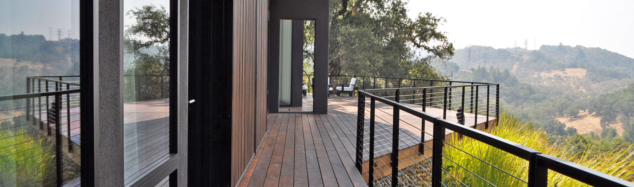 A modern outdoor balcony with wooden flooring and black metal railing overlooking a hilly landscape with trees and distant hills.