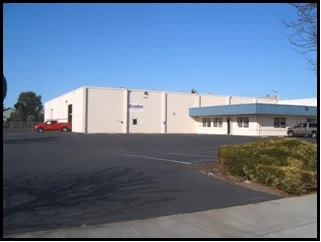 A large beige commercial building with a flat roof, white sides, and a small parking lot with a red pickup truck and a gray car parked outside, under a clear blue sky.
