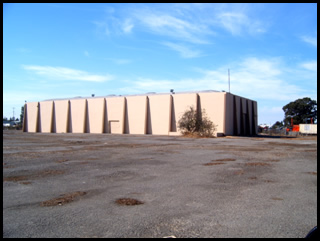 Large beige building with vertical structural elements, set in an open paved area with a clear blue sky.