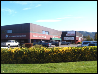 Parking lot in front of a commercial building with cars parked, surrounded by yellow flowers and green grass, under a blue sky.