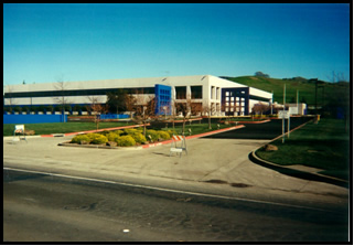 A modern multi-story building with large windows, surrounded by landscaped lawns, trees, and parking lot under a clear blue sky.