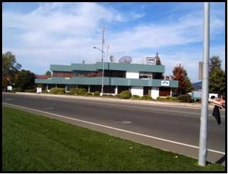 A modern, light blue and gray multi-story building with antennas and satellite dishes on the roof, located along a busy street with a grassy median and some trees in the background.
