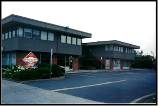 A two-story commercial building with a black exterior, large windows, and a parking lot in front, along with a small sign near the entrance.