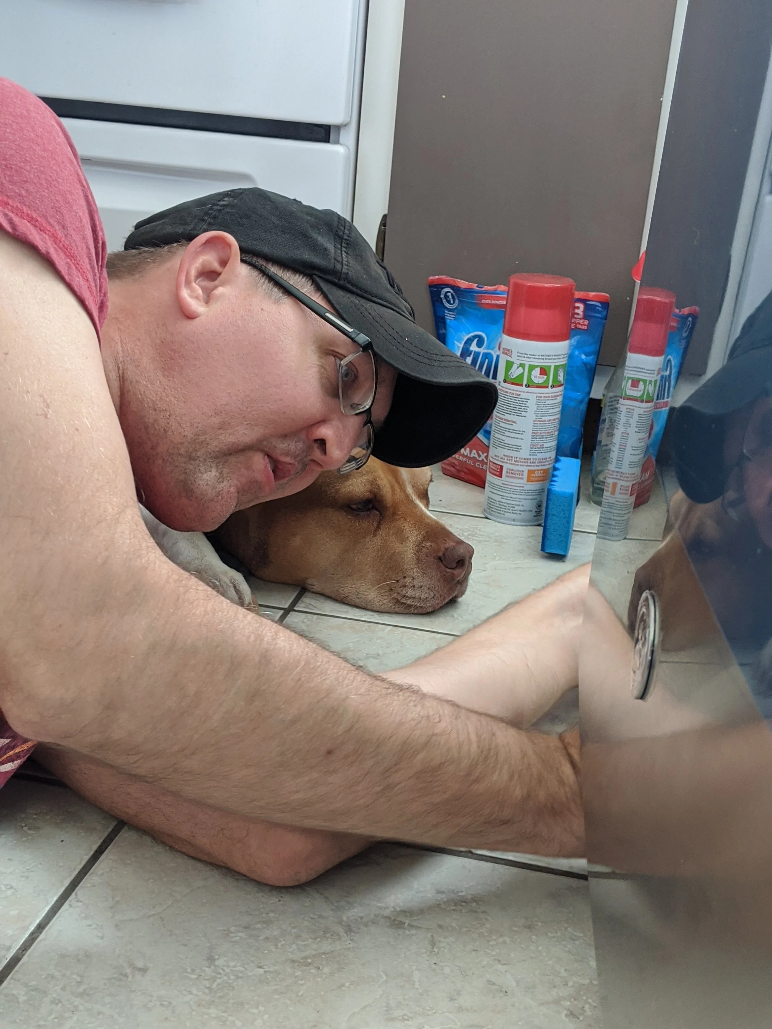 A man lying on the kitchen floor resting his head on a large dog. The man wears glasses, a black cap, and a red shirt. The dog has a brown coat. Several cans and bags of cleaning products and fabric softener are on the floor behind them. Some of these products have red caps and labels with green and red instructions.