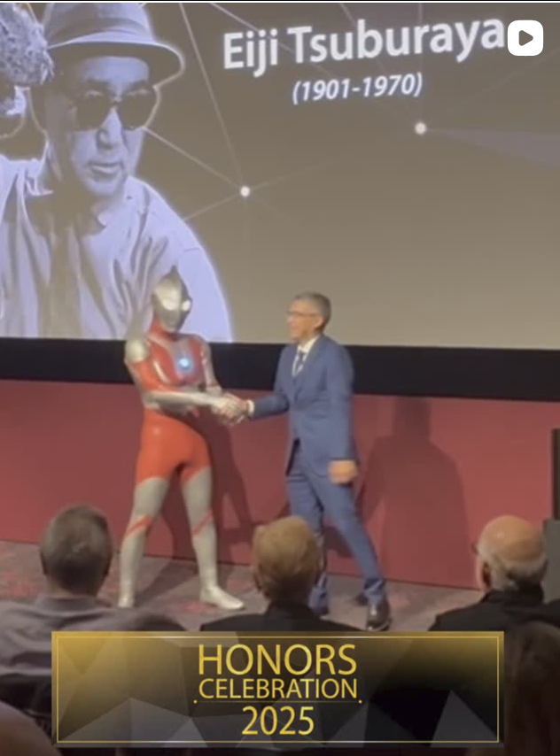 A man in a suit shaking hands with a person dressed as Iron Man at the 2025 Honors Celebration event, with a large screen in the background displaying a photo and information about Eiji Tsuburaya (1901-1970).