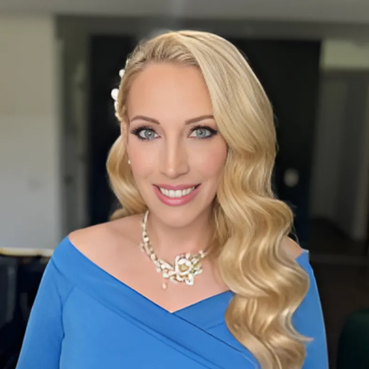 A woman with long blonde hair styled in loose waves, wearing a blue off-the-shoulder dress and a pearl necklace, smiling at the camera in an indoor setting.