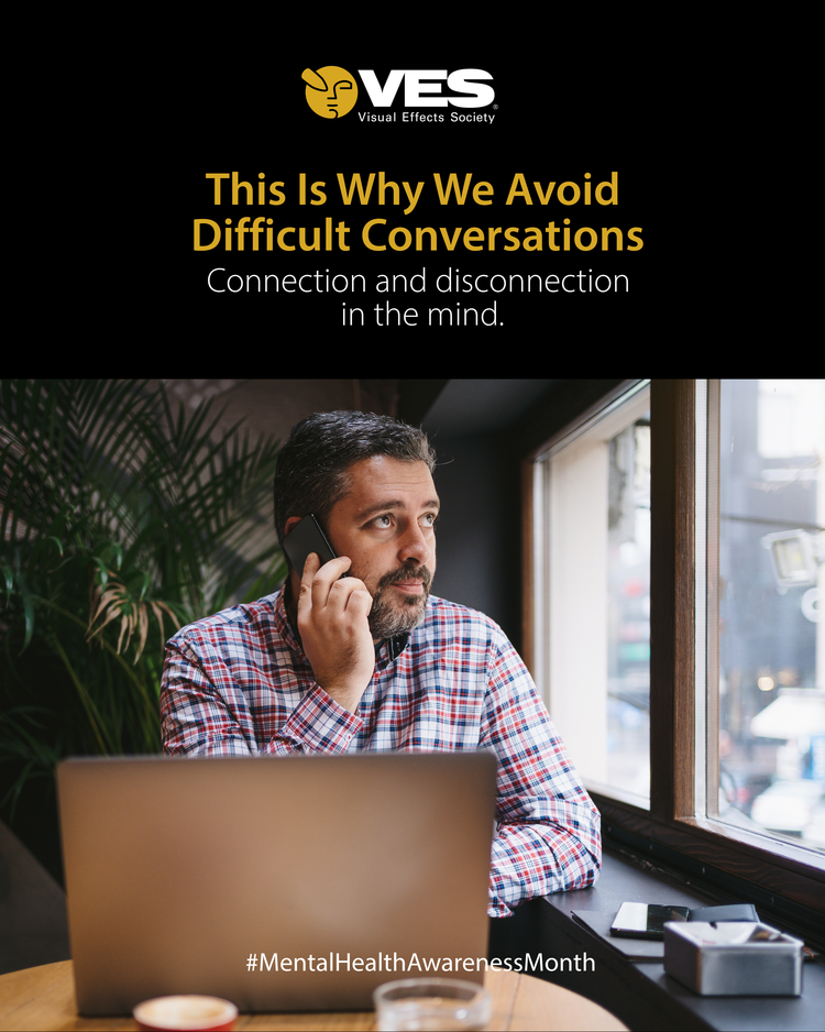 A man with a beard and checked shirt sitting at a table, holding a phone to his ear, with a laptop in front of him near a window, signifying mental health awareness.