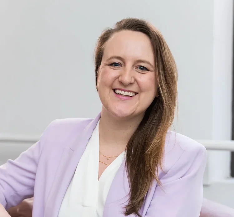 A smiling woman with long brown hair, wearing a white blouse and a light purple blazer, sitting in an office setting.