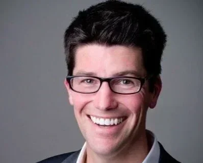 A smiling man with dark hair, glasses, and a suit, against a plain background.