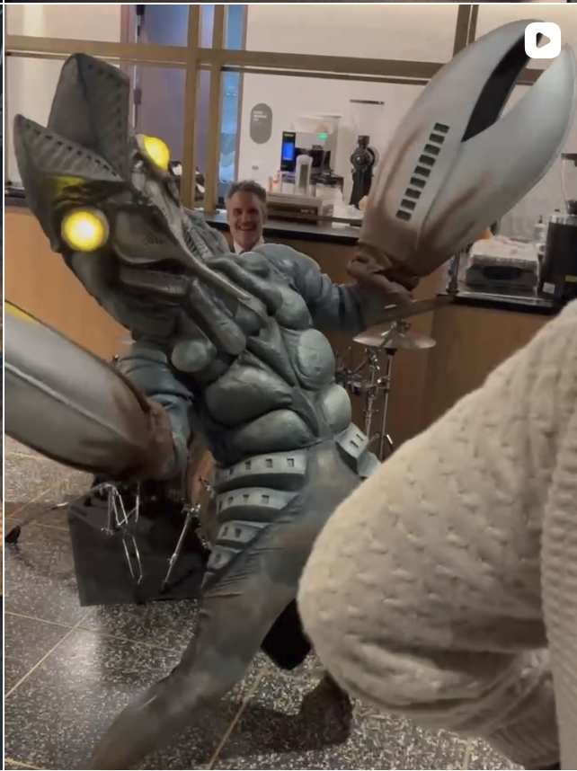 A person in a robotic suit taking a selfie in a cafe, with a happy woman smiling in the background.
