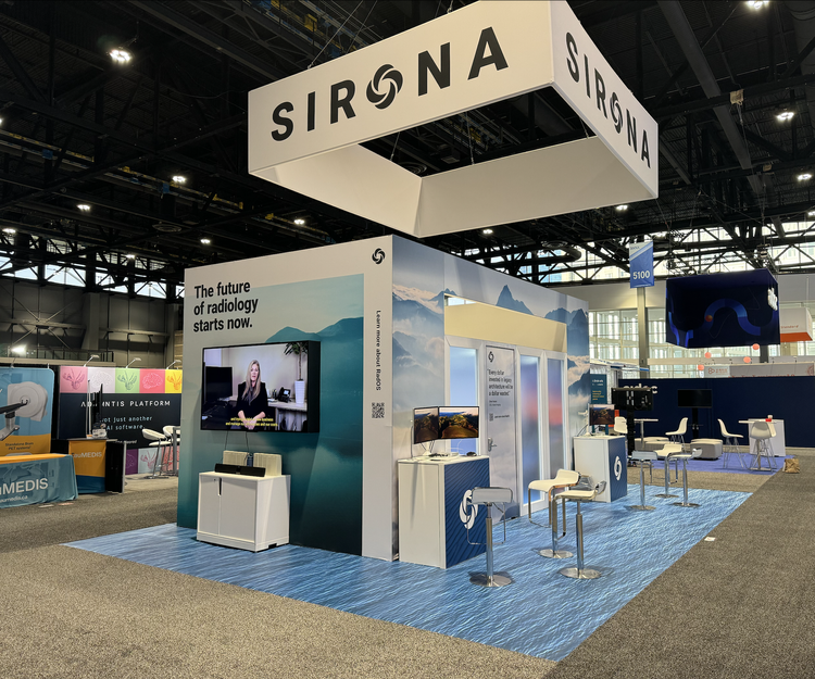 Trade show booth for Sirona, a radiology company, with a large hanging sign displaying the company name. The booth features a wall with the slogan, "The future of radiology starts now," a monitor with a woman speaking, and several smaller screens and chairs for demonstrations or discussions. The area has a blue carpet and is situated inside a large exhibition hall.