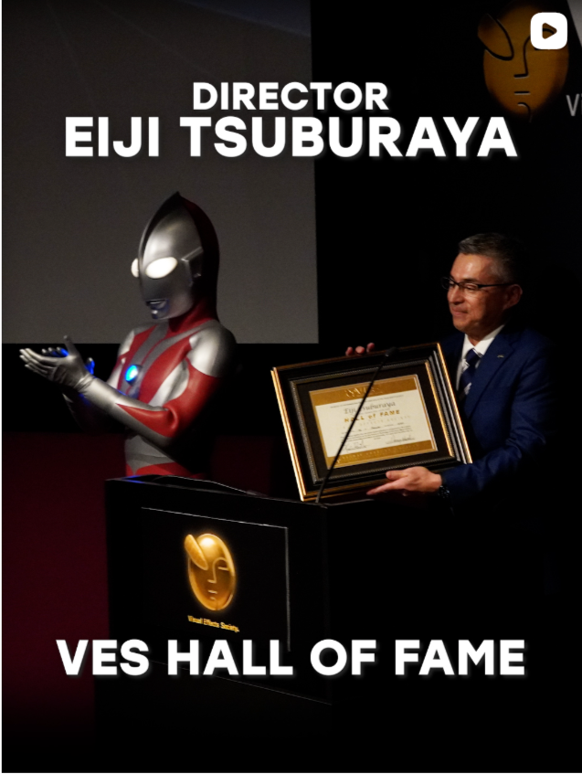 A man in a suit receiving a framed award at the VES Hall of Fame event, standing beside a robot with a humanoid appearance and a glowing emblem on its chest, in a dimly lit room with a screen displaying the words 'Director Eiji Tsuburaya'.