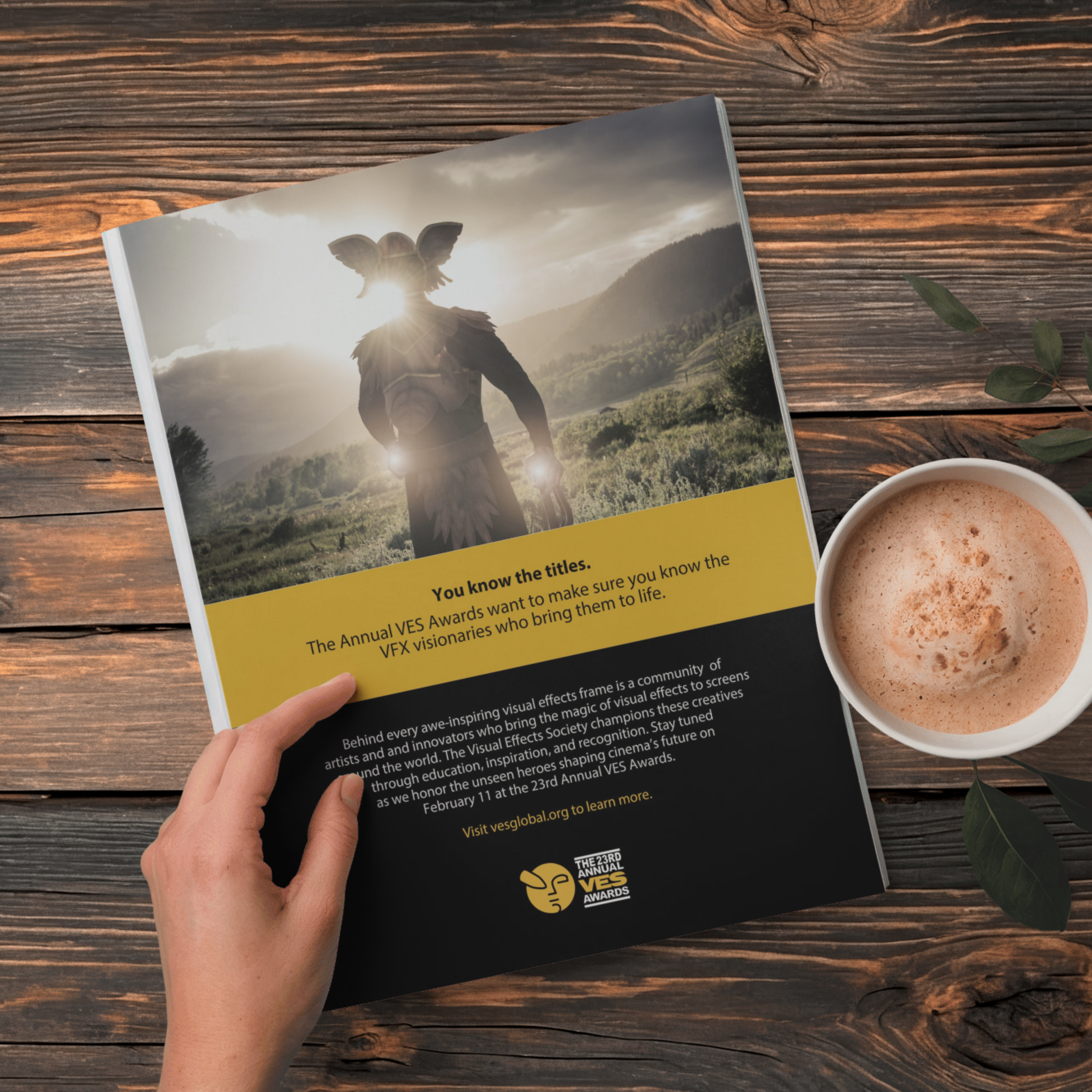 A person's hand holding a flyer with a background photo of a person wearing a headdress resembling a butterfly, standing outdoors with mountains and cloudy sky in the background. The flyer discusses the 23rd Annual VES Awards and visual effects community.