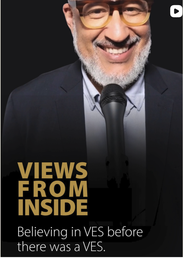 Smiling man with glasses and a beard, holding a microphone, wearing a suit and standing against a black background.