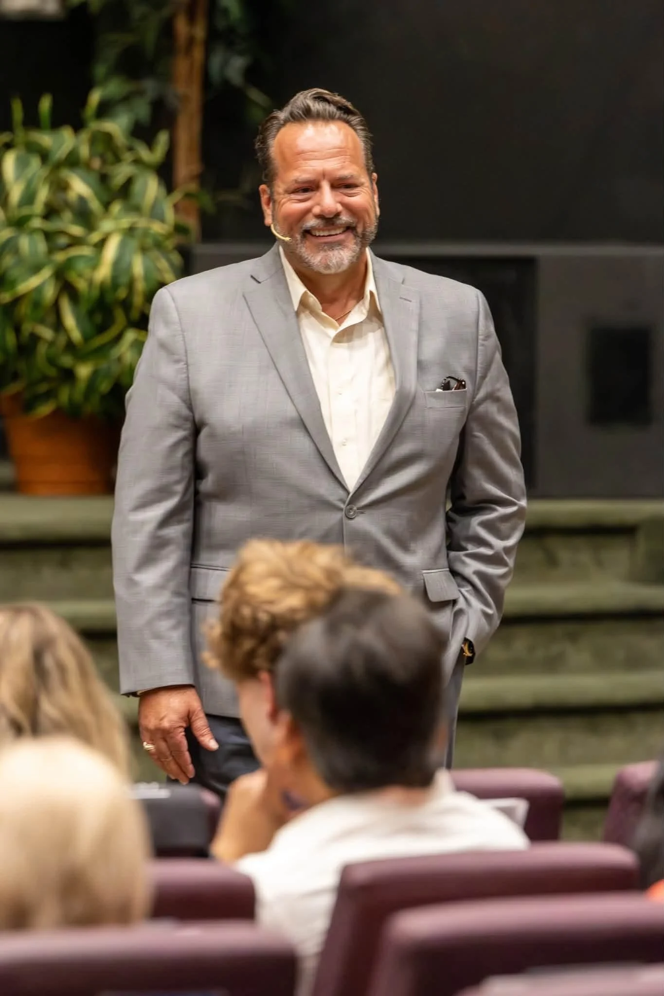 A smiling man in a gray suit with a beard and mustache, wearing a wireless microphone, standing in front of an audience in a room with dark walls and large green plants.