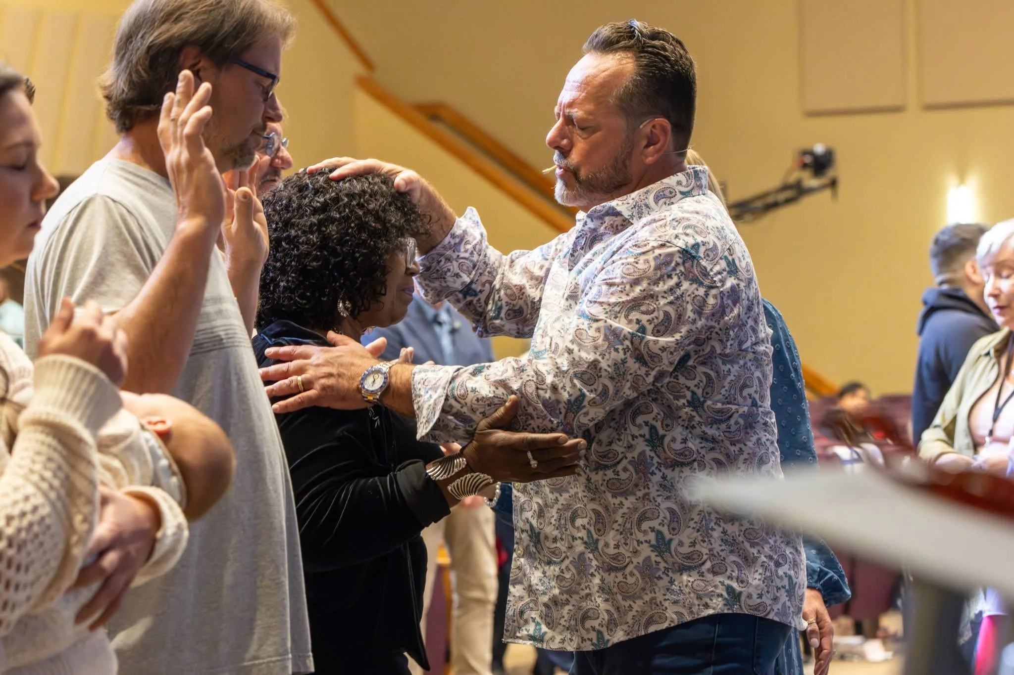 A man with a patterned shirt is praying over and laying hands on a woman in a black jacket, with other people praying with eyes closed and hands raised nearby.