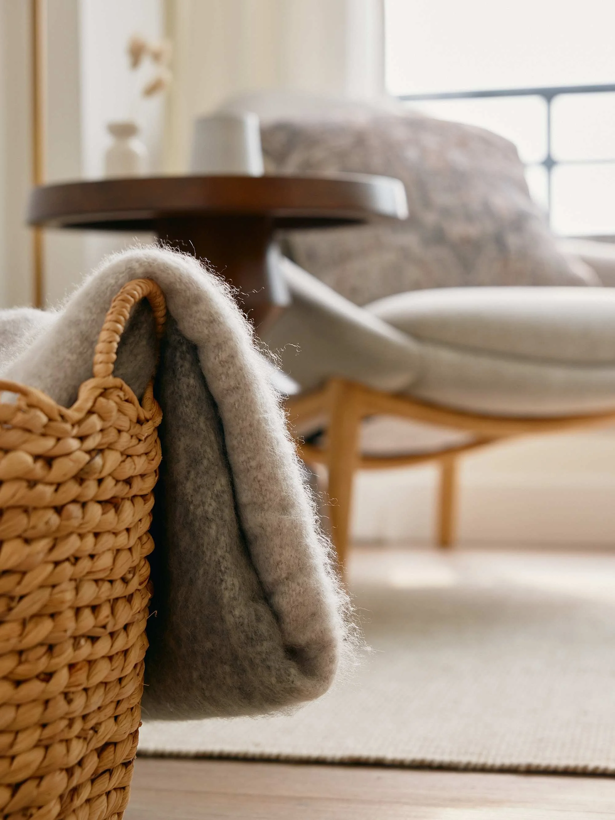 Close-up of a woven basket and a fuzzy gray blanket in the foreground, a wooden table and a cozy armchair with a cushion near a window in the background.