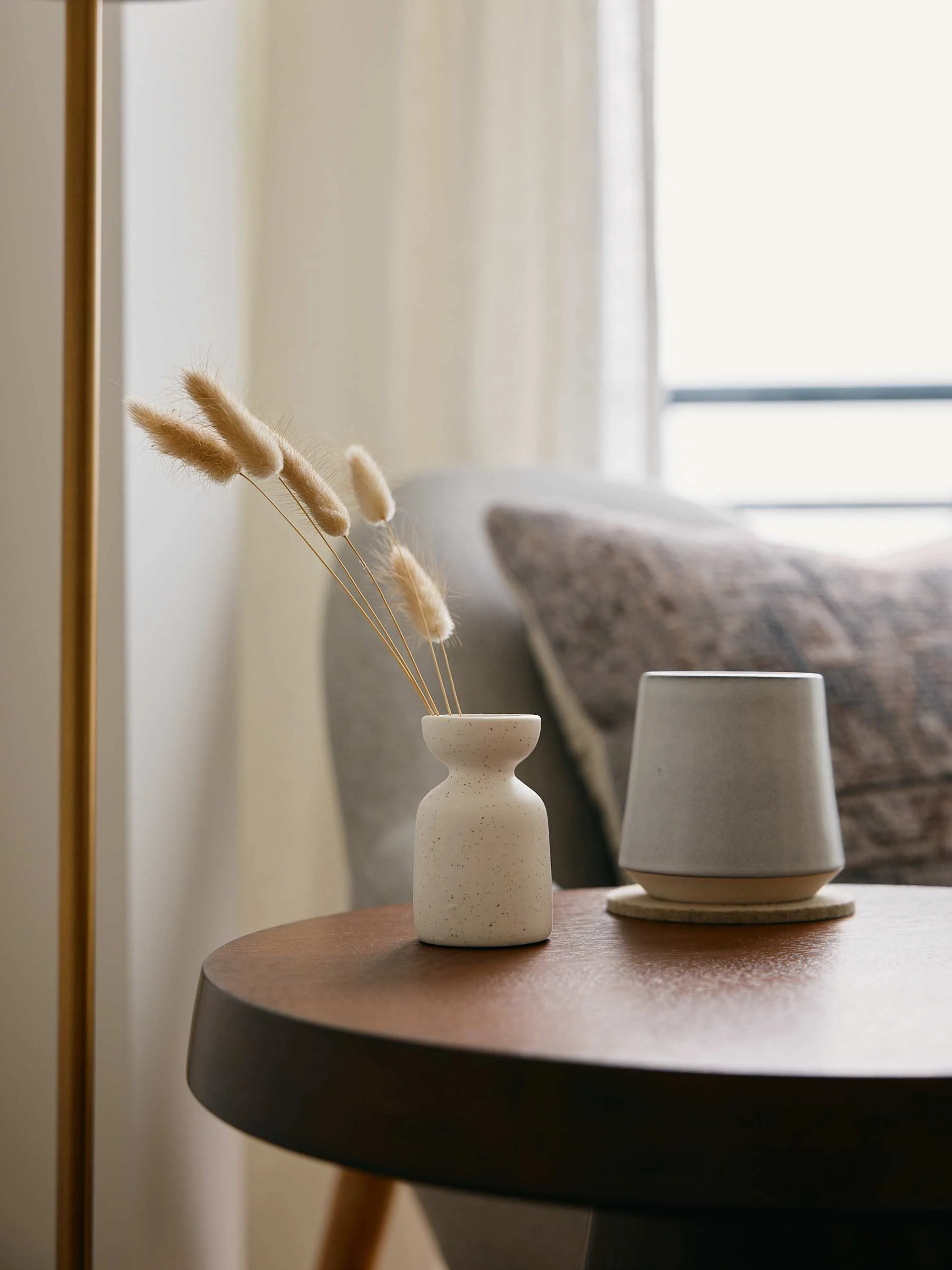 A beige ceramic vase with a small neck holding dried bunny tail grass stems, placed on a round wooden table. In the background, a gray ceramic lamp sits on the table, and a sofa with a patterned pillow is visible near a window with white curtains.