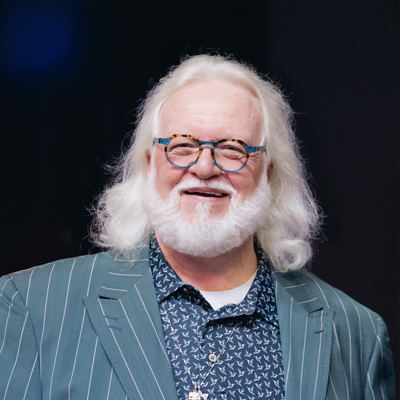 A smiling elderly man with long white hair and a beard, wearing glasses, a blue patterned shirt, and a pinstriped blazer.