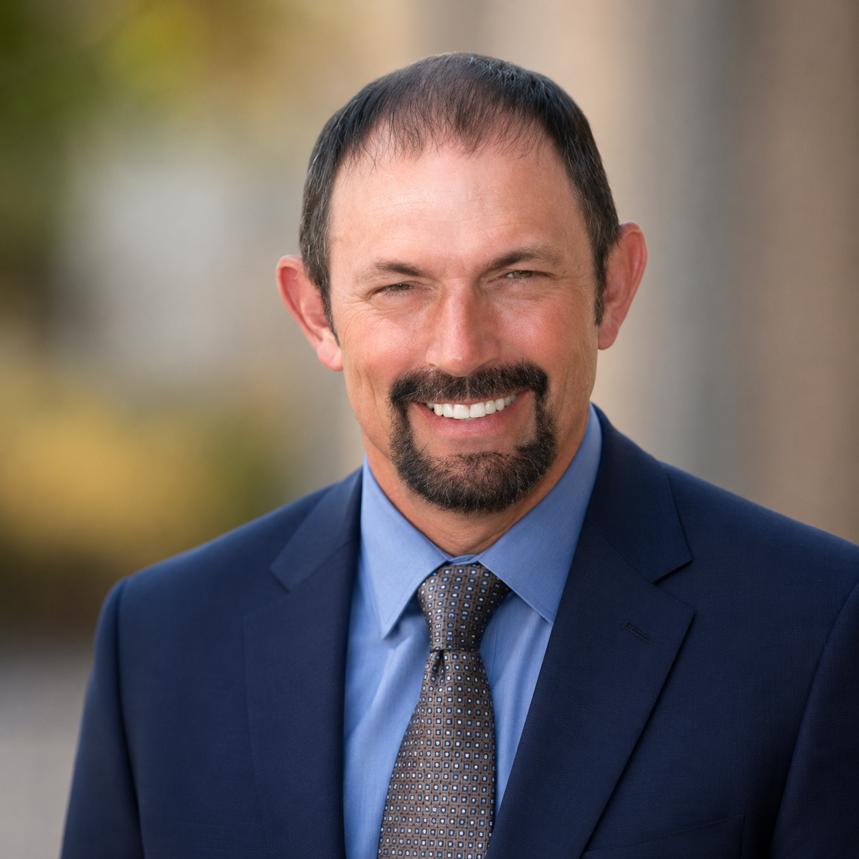 A smiling man in a suit with a blue shirt and patterned tie, outdoors with blurred greenery in the background.