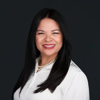 Headshot of a woman with long black hair, wearing a white blouse and pearl necklace, smiling against a dark background.