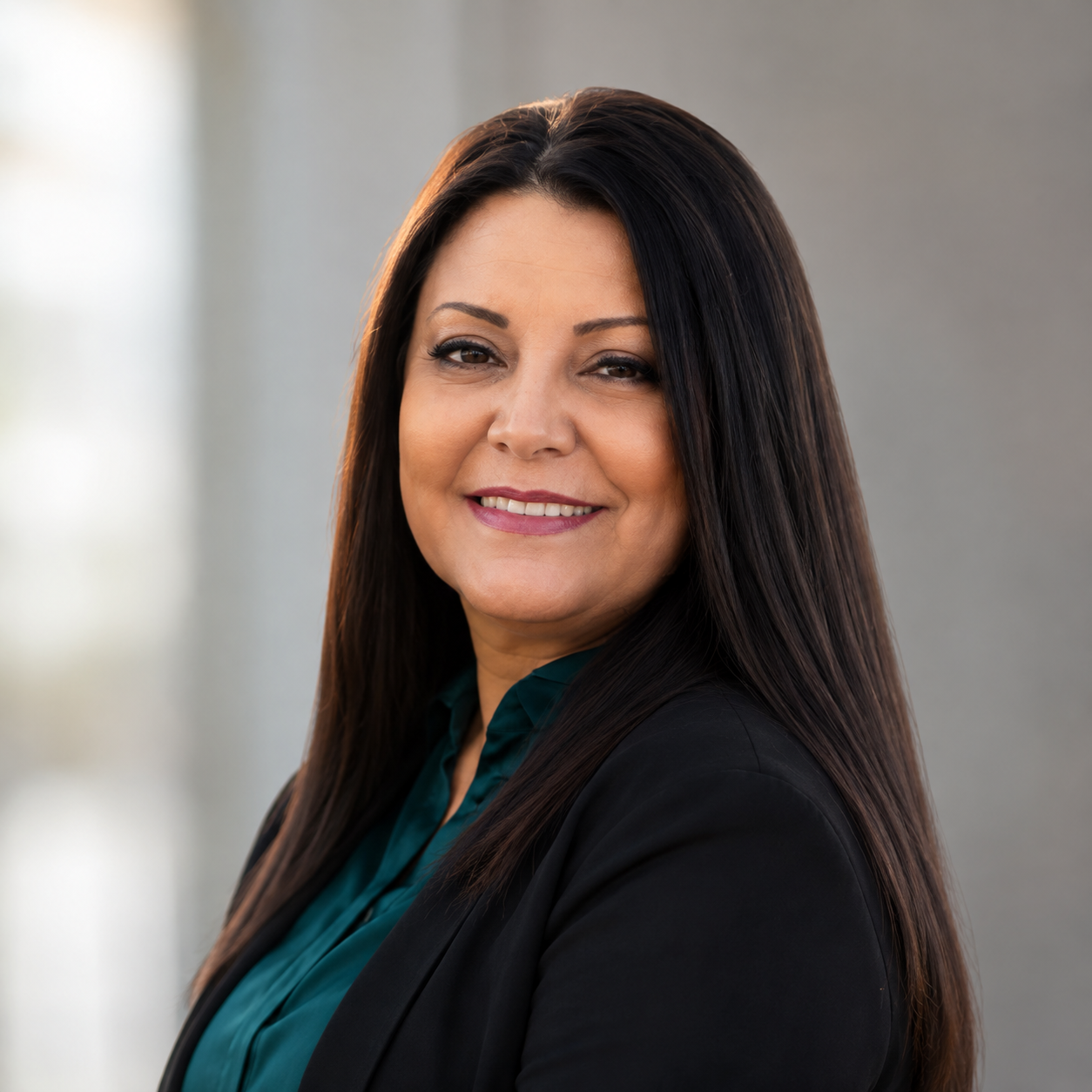 Professional woman with long dark hair smiling, wearing a black blazer and teal blouse, standing in an office setting.