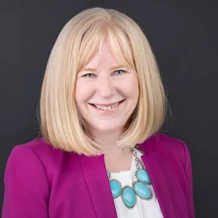 Portrait of a smiling woman with blonde hair wearing a purple blazer and a statement necklace, against a dark background.