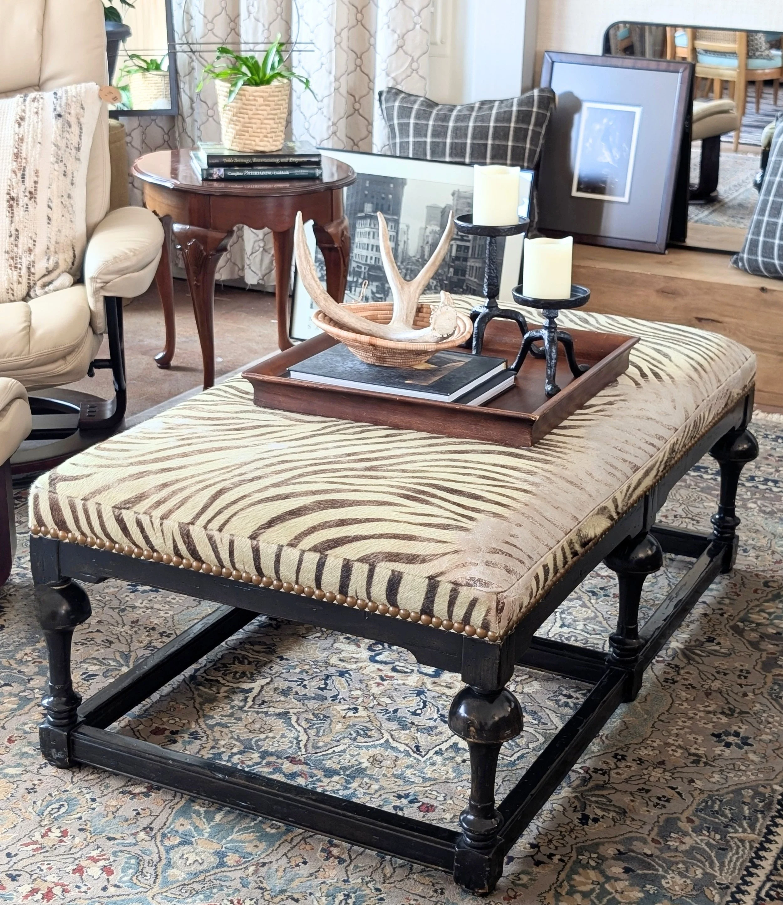 Living room with a vintage-style black wooden bench with cushioned upholstery featuring zebra print, placed on a patterned rug. The bench is decorated with a wooden tray containing a faux antler and books. In the background, there's a round wooden side table with a potted plant, an ornate mirror leaning against the wall, a plaid pillow, framed art pieces, and various furniture including a beige armchair and a dining table with chairs.