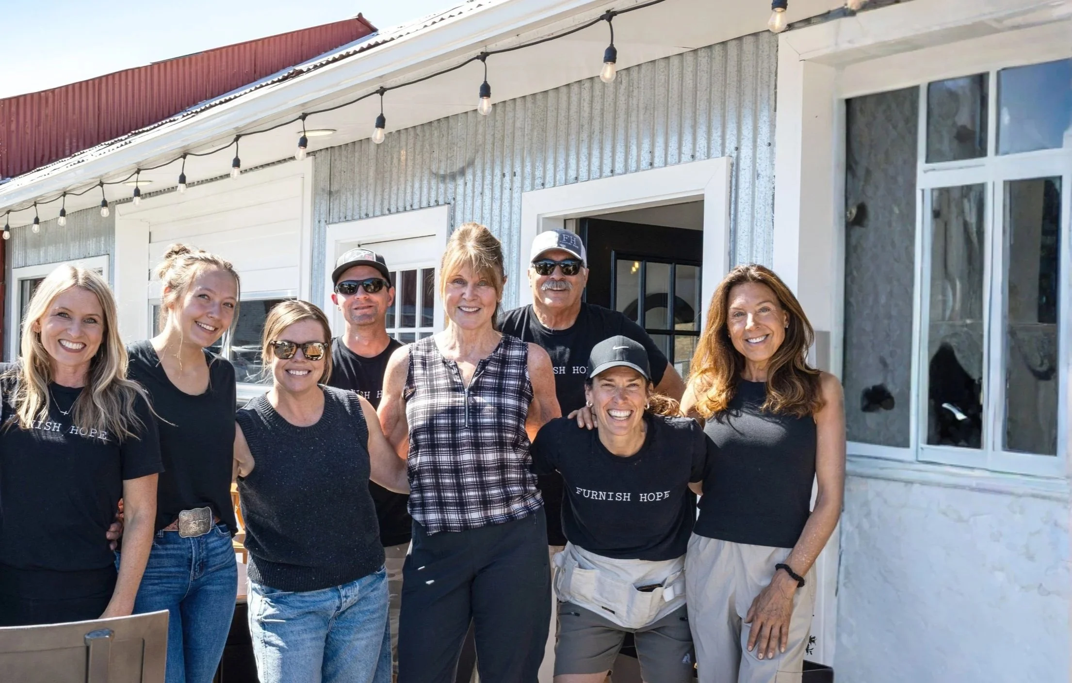 Group of nine smiling people standing outside a building with string lights on the roof.