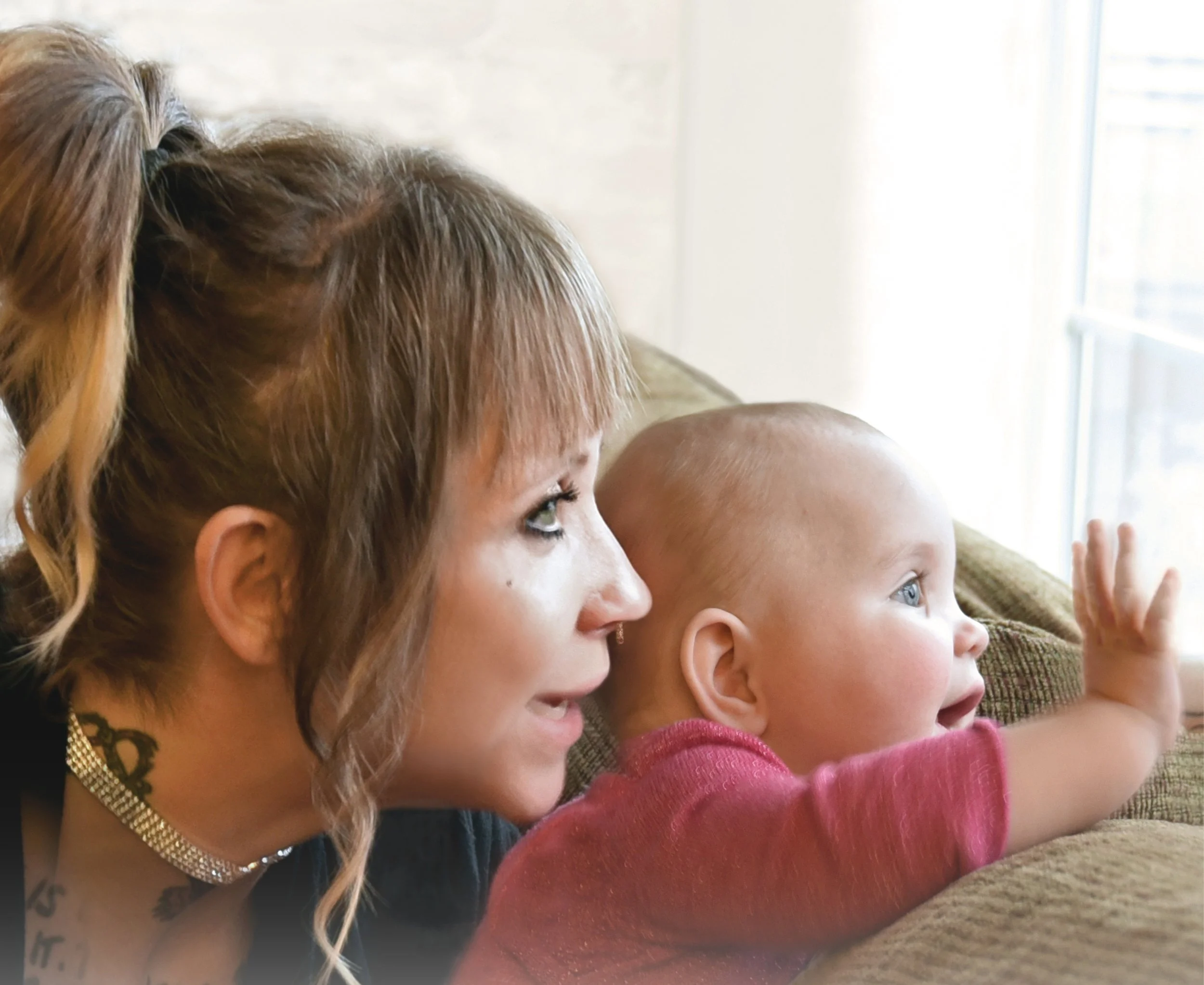 Close-up image of a woman with reddish hair, and two children, all facing a window. The woman is wearing a necklace and has face paint on her cheek. One child is in a pink shirt and the other is bald, with their hands reaching out towards the window.