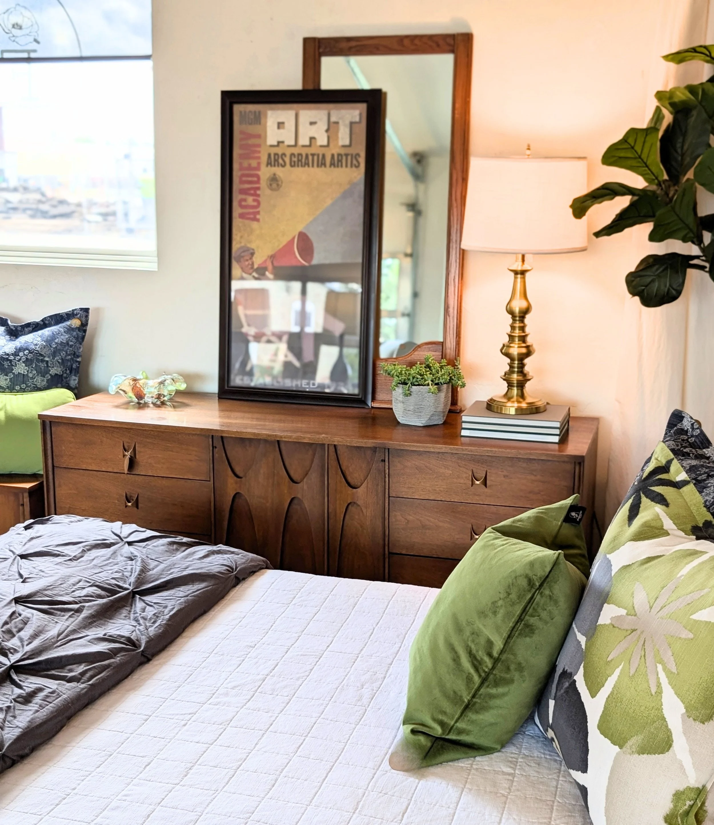 A bedroom featuring a neatly made bed with a white quilt, green and patterned pillows, a wooden dresser with a large mirror, a framed artistic poster, a table lamp with a brass base, a potted plant, and a window with blinds.
