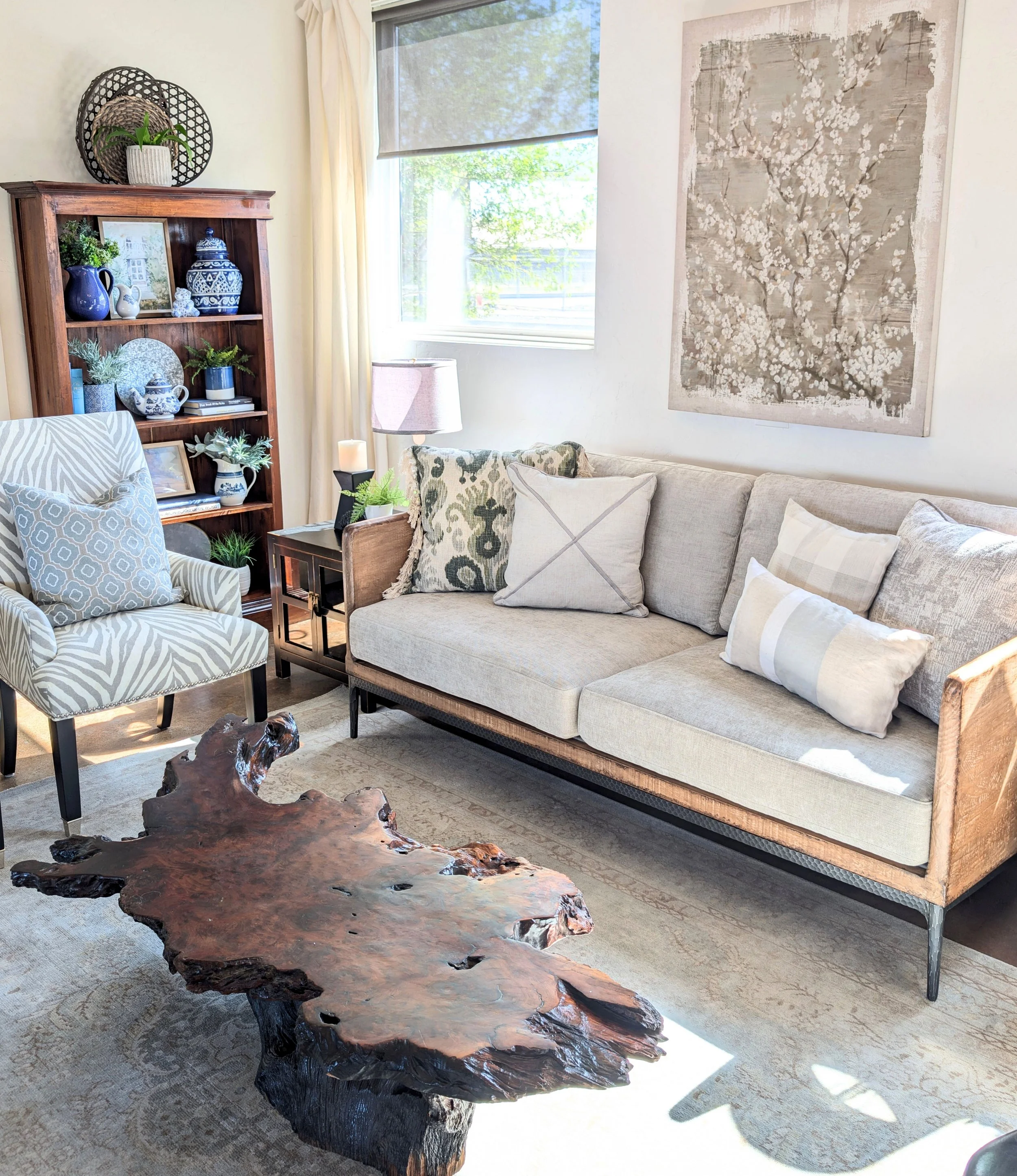 Living room with beige sofa and decorative pillows, wooden coffee table, wooden bookshelf with blue and white decorative items, large wall art, window with light-colored curtains, and patterned armchair.