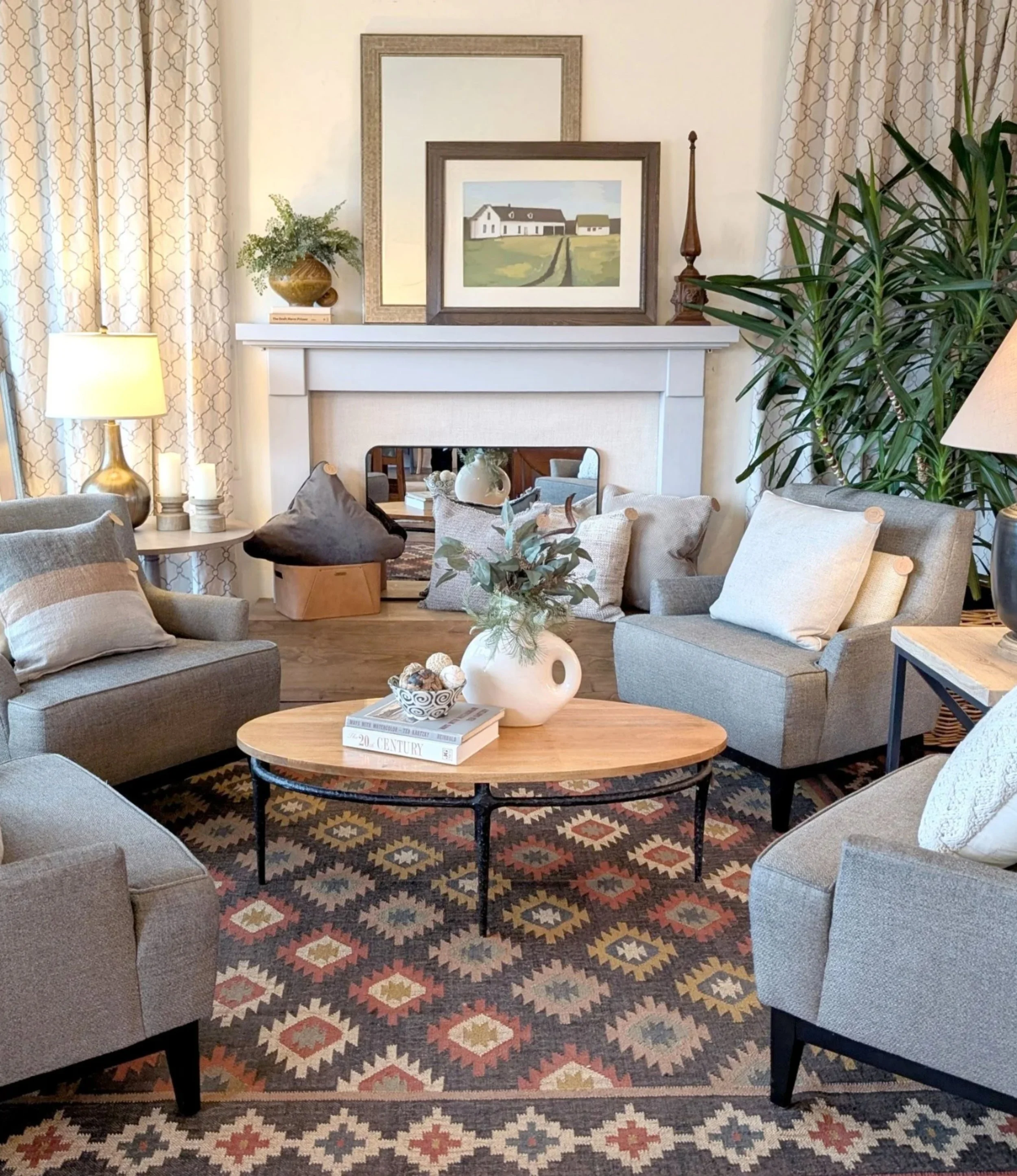 Living room with patterned area rug, gray sofas with pillows, wooden coffee table with decorative bowl and books, fireplace mantel with framed pictures and decor, large house painting, potted plant, table lamp, and curtains.