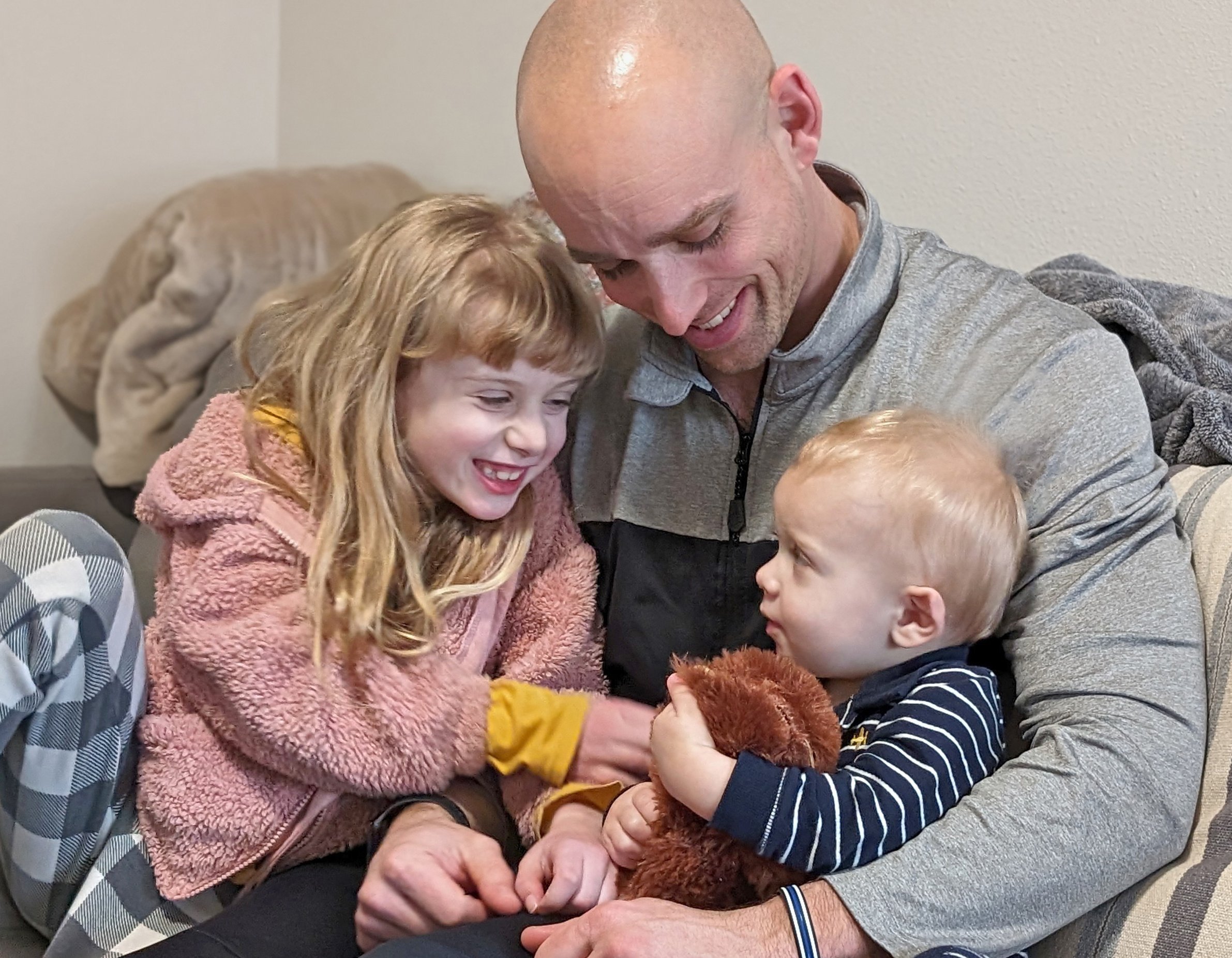A man and two children, a girl with long red hair and a boy with blond hair, are sitting close together, smiling and looking at the boy. They are sharing a joyful moment inside a room with a beige wall and some clothing in the background.
