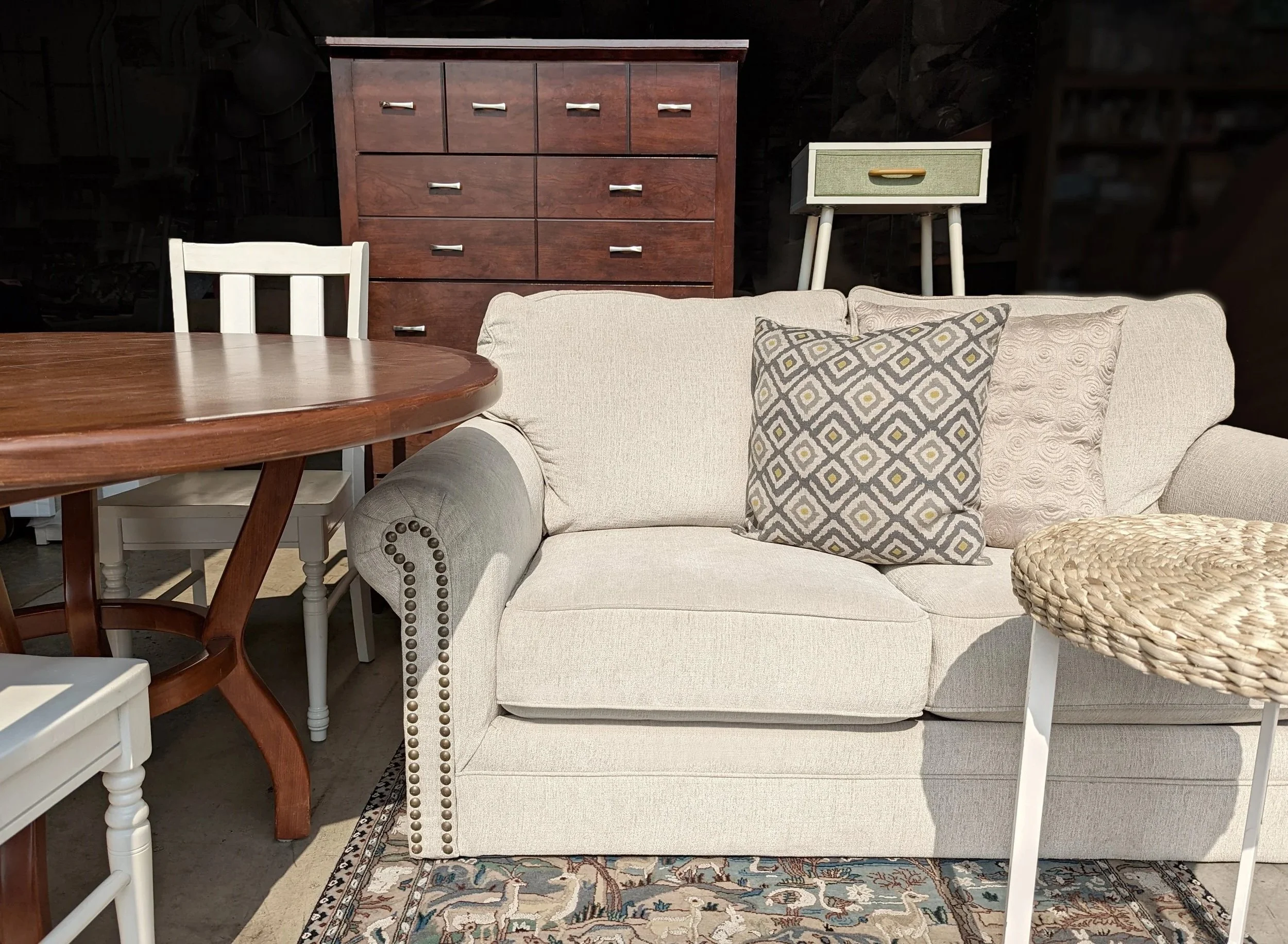 A beige upholstered sofa with two patterned throw pillows, a round woven side table, a wooden round dining table with a white chair, a dark wooden dresser, and a small white and green nightstand against a dark background.