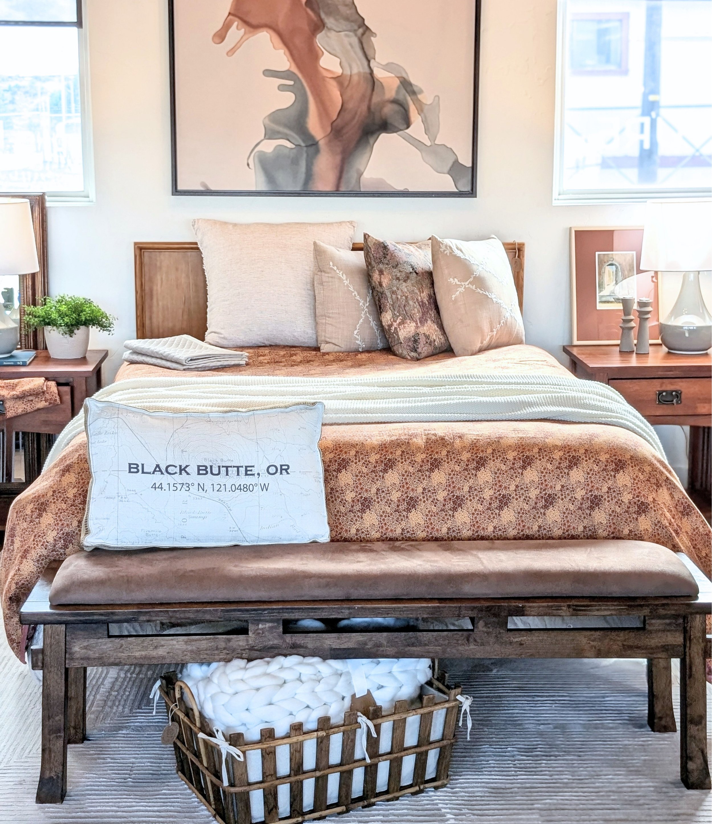 A bed with a wooden headboard, covered with a beige bedspread and several decorative pillows, in a bright room with large windows. Two nightstands with lamps are on either side of the bed. A large abstract painting hangs on the wall behind the bed. A laundry basket filled with white items is placed at the foot of the bed.